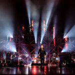 Bald man wearing all black stands center stage with bright lights shooting rays of white toward the sky around him. He is walking toward the camera. His mandates playing electric guitar, bass, and drums are in the background, on stage playing.