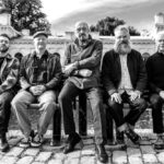 Five men sit togethers on a bench in front of an ornate white building. From left, there is a younger man in a flannel shirt and demin jeans. He has a small beard and boots on. The next man has a newsboy hat on and his hands clasped in front of him and his button-up shirt. He has sneakers on. The next man, the center of the group, is an older bald man with a sweatshirt zipped up, pants, and sneakers. He has glasses and a dark goatee. The next man has a long grey beard and spiky grey hair. He is wearing a grey jacket over a checkered shirt with denim jeans and sneakers. The last man the furthest to the right is wearing a solid black shirt buttoned up high and denim jeans and sneakers. He has short white hair. The photo is in black and white.