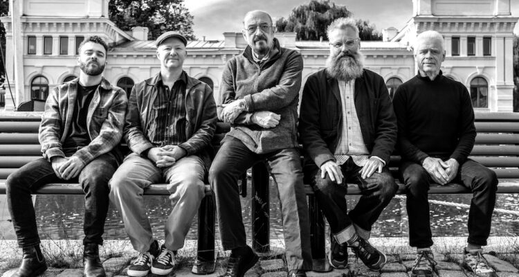 Five men sit togethers on a bench in front of an ornate white building. From left, there is a younger man in a flannel shirt and demin jeans. He has a small beard and boots on. The next man has a newsboy hat on and his hands clasped in front of him and his button-up shirt. He has sneakers on. The next man, the center of the group, is an older bald man with a sweatshirt zipped up, pants, and sneakers. He has glasses and a dark goatee. The next man has a long grey beard and spiky grey hair. He is wearing a grey jacket over a checkered shirt with denim jeans and sneakers. The last man the furthest to the right is wearing a solid black shirt buttoned up high and denim jeans and sneakers. He has short white hair. The photo is in black and white.