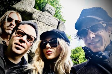 Four adults take a selfie in front of rocks and greenery. From left is a blonde man is sunglasses on, a dark-haired man with glasses on, a blonde woman with a black conductor hat on and black sunglasses, and a man with sunglasses on, a polka dot scarf under a jacket, and a striped conductor hat. The last man is the only one with a smile.