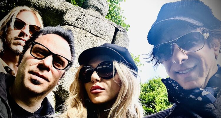 Four adults take a selfie in front of rocks and greenery. From left is a blonde man is sunglasses on, a dark-haired man with glasses on, a blonde woman with a black conductor hat on and black sunglasses, and a man with sunglasses on, a polka dot scarf under a jacket, and a striped conductor hat. The last man is the only one with a smile.