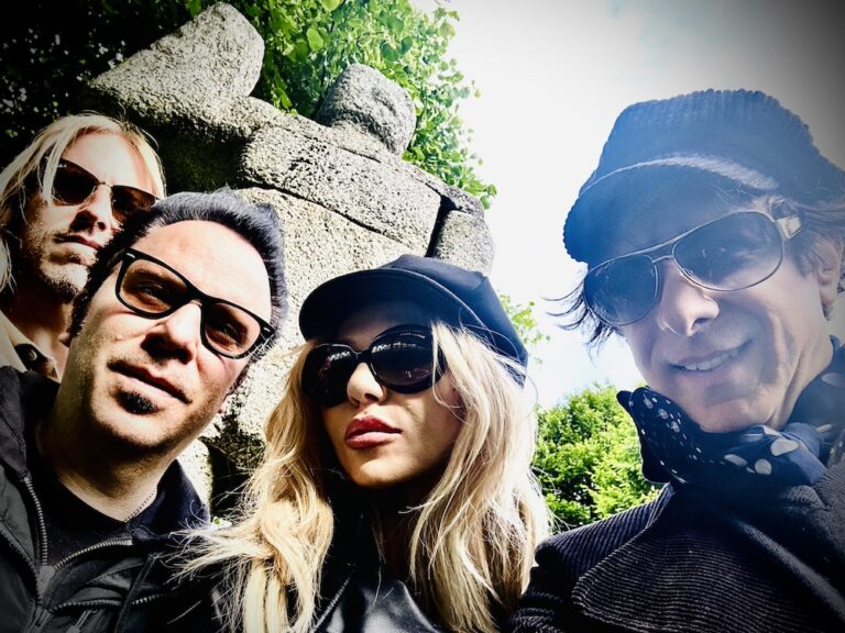 Four adults take a selfie in front of rocks and greenery. From left is a blonde man is sunglasses on, a dark-haired man with glasses on, a blonde woman with a black conductor hat on and black sunglasses, and a man with sunglasses on, a polka dot scarf under a jacket, and a striped conductor hat. The last man is the only one with a smile.