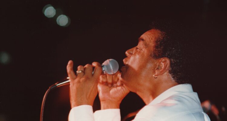 An African-American man sings into a microphone. The background is dark. His eyes are closed and the hand holding the microphone has a wedding ring on his ring finger. He has a small hoop earring and is wearing a white shirt.